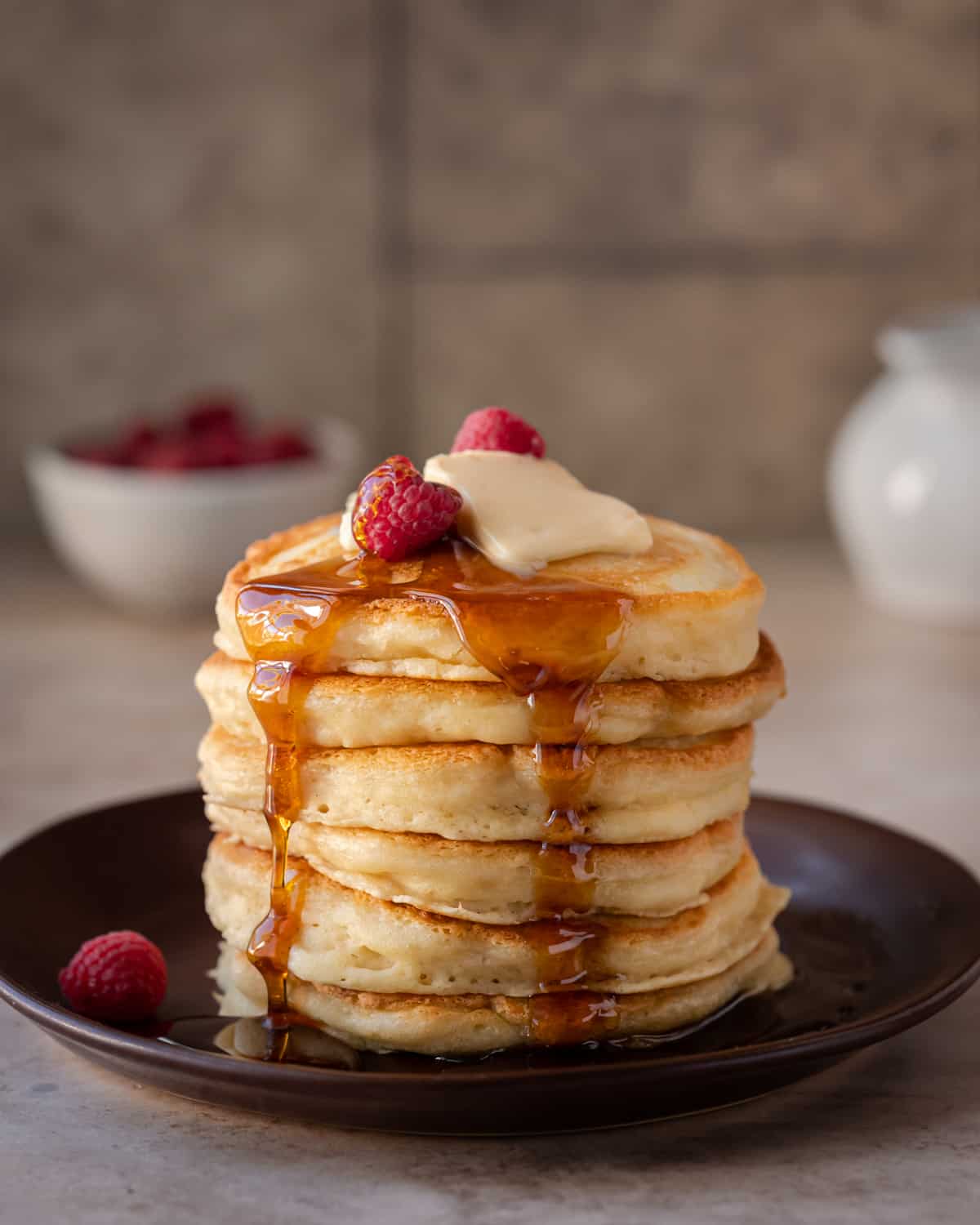 woman's hands pouring maple syrup onto a stack of fluffy golden vegan pancakes on a brown plate.